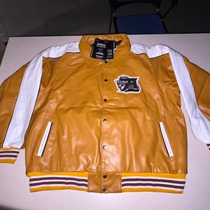 May include: A mustard yellow jacket with white stripes running down the sleeves. The jacket features a collar, a front button closure, and a black and white patch on the chest. The cuffs and hem are ribbed with mustard, white, and dark brown stripes.