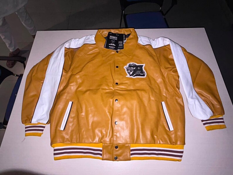 May include: A mustard yellow jacket with white stripes running down the sleeves. The jacket features a collar, a front button closure, and a black and white patch on the chest. The cuffs and hem are ribbed with mustard, white, and dark brown stripes.