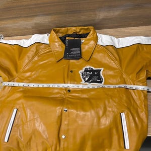 May include: A mustard yellow jacket with white shoulder panels and black piping. It features a black and white embroidered patch on the chest, silver snap buttons, and two front pockets. A measuring tape across the chest shows a width of 31 inches.