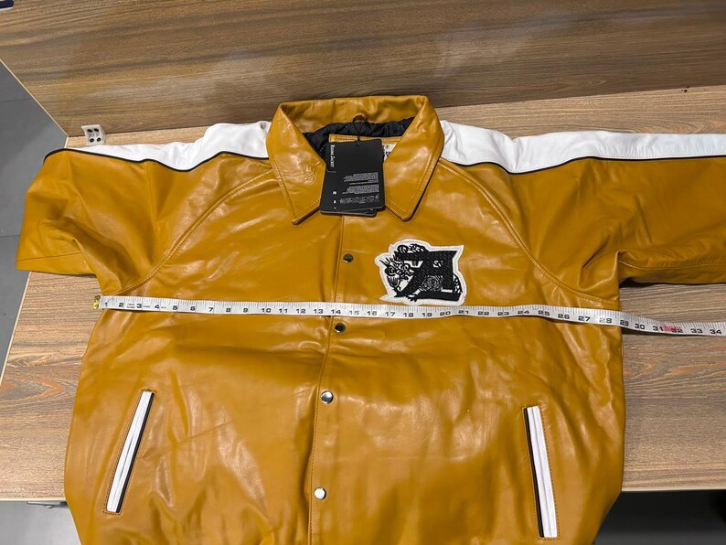 May include: A mustard yellow jacket with white shoulder panels and black piping. It features a black and white embroidered patch on the chest, silver snap buttons, and two front pockets. A measuring tape across the chest shows a width of 31 inches.