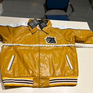 May include: A mustard yellow varsity jacket with white shoulder stripes, a pointed collar, and snap button closures. A black and white patch is on the chest. A measuring tape across the chest shows a width of approximately 25 inches.