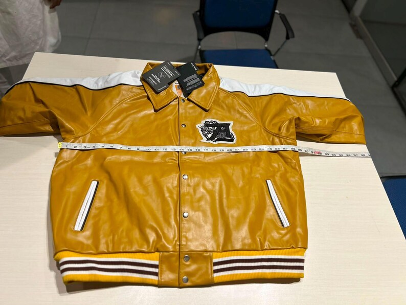 May include: A mustard yellow varsity jacket with white shoulder stripes, a pointed collar, and snap button closures. A black and white patch is on the chest. A measuring tape across the chest shows a width of approximately 25 inches.