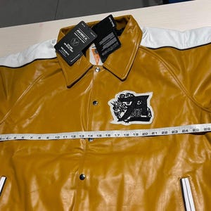 May include: A mustard yellow jacket with white shoulder panels, a black and white embroidered patch on the chest, and silver snap buttons. A white measuring tape is placed across the front, showing a width measurement of approximately 28 inches.