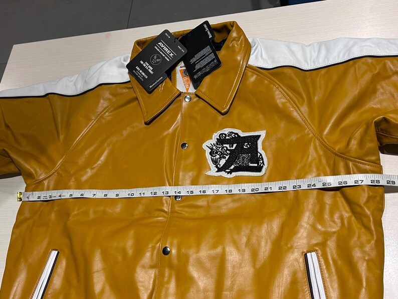 May include: A mustard yellow jacket with white shoulder panels, a black and white embroidered patch on the chest, and silver snap buttons. A white measuring tape is placed across the front, showing a width measurement of approximately 28 inches.