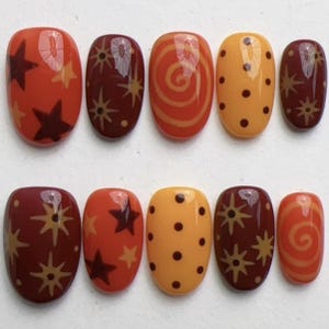 May include: A set of ten press-on nails featuring a glossy finish in shades of orange, yellow, and deep red. The designs include dark brown stars, light yellow starbursts, orange spirals, and dark brown polka dots on various nail surfaces.