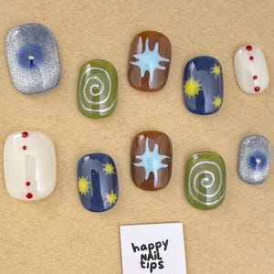 Könnte beinhalten: Eine Sammlung von zehn künstlichen Fingernägeln mit verschiedenen Designs. Die Nägel zeigen Farben wie Blau, Grün, Braun und Weiß, mit Mustern wie Sternen, Spiralen und Punkten. Der Text "happy NAIL tips" ist sichtbar.