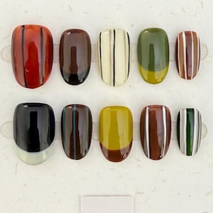 Puede incluir: Un juego de diez uñas postizas con diversos diseños, incluyendo rayas verticales en marrón, blanco y negro, bloques sólidos en verde oliva, amarillo mostaza y negro intenso, y patrones de bloques de color con acabados brillantes.
