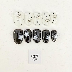 May include: Two sets of oval-shaped press-on nails. One set is white with black polka dots and silver star embellishments. The other is black with silver star accents. The nails are displayed on clear plastic holders. A small white square reads "happy nail tips".