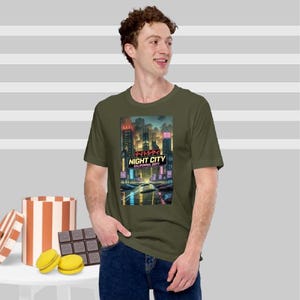 Op de afbeelding: Olijfgroen t-shirt met een levendige afbeelding van een futuristische stad met de tekst "NIGHT CITY CALIFORNIA 2077". Het ontwerp toont neonlichten en hoge gebouwen. Het t-shirt wordt door een persoon gedragen.