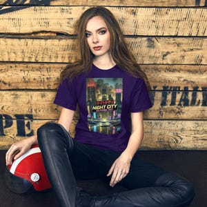 Op de afbeelding: Een paars t-shirt met een futuristisch stadslandschapontwerp, inclusief de tekst "NIGHT CITY CALIFORNIA 2077" in het Engels en Japans. Het ontwerp toont een neonverlicht stadslandschap. Een rood-witte helm ligt op de grond.