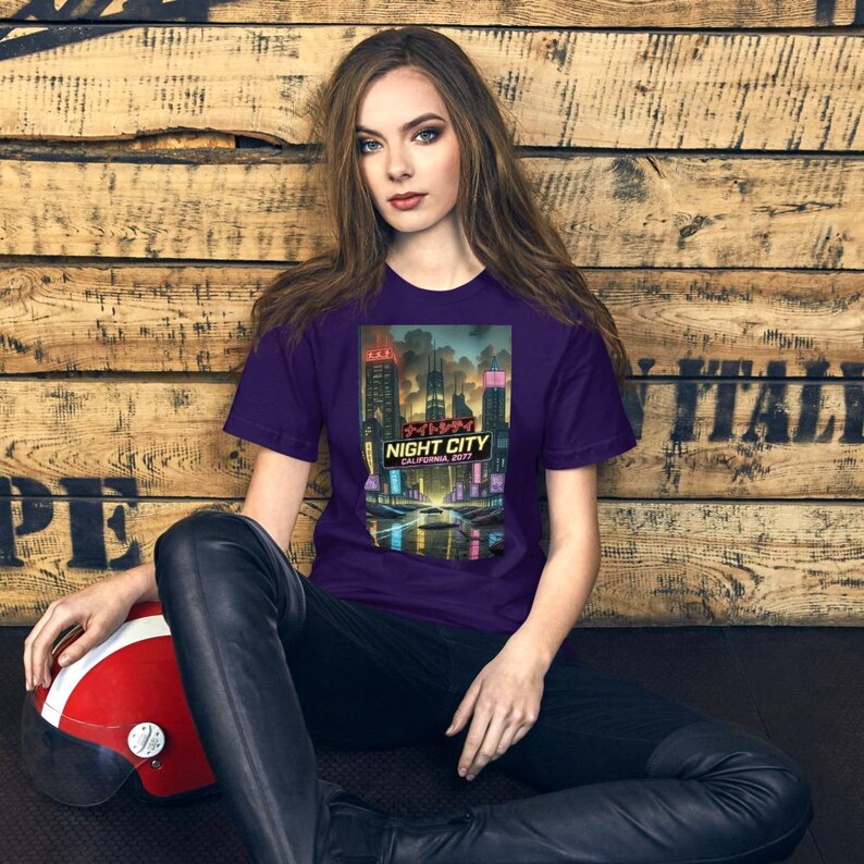 Op de afbeelding: Een paars t-shirt met een futuristisch stadslandschapontwerp, inclusief de tekst "NIGHT CITY CALIFORNIA 2077" in het Engels en Japans. Het ontwerp toont een neonverlicht stadslandschap. Een rood-witte helm ligt op de grond.