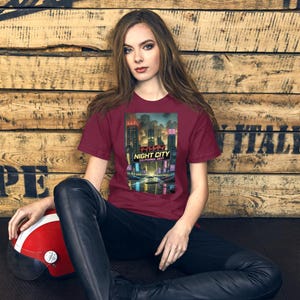 Op de afbeelding: Een bordeauxrood t-shirt met een futuristische stadsafbeelding en de tekst "NIGHT CITY CALIFORNIA 2077" in het Engels en Japans. Het t-shirt wordt door een persoon gedragen.