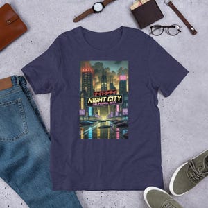 Op de afbeelding: Marineblauw T-shirt met een kleurrijke afbeelding van een futuristische stad, met neonlichten en de tekst "NIGHT CITY CALIFORNIA 2077". Het ontwerp bevat Japanse tekst en een waterreflectie.