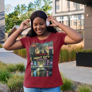 Op de afbeelding: Bordeauxrood t-shirt met een afbeelding van een futuristische stadsgezicht 's nachts, met de tekst "NIGHT CITY CALIFORNIA, 2077". Het shirt wordt gecombineerd met een blauwe spijkerbroek. De persoon draagt een witte koptelefoon.