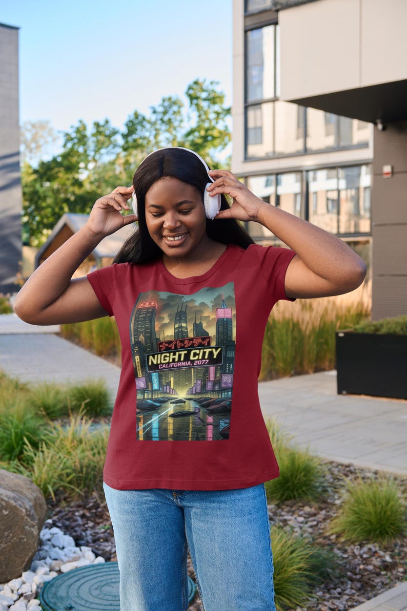 Op de afbeelding: Bordeauxrood t-shirt met een afbeelding van een futuristische stadsgezicht 's nachts, met de tekst "NIGHT CITY CALIFORNIA, 2077". Het shirt wordt gecombineerd met een blauwe spijkerbroek. De persoon draagt een witte koptelefoon.