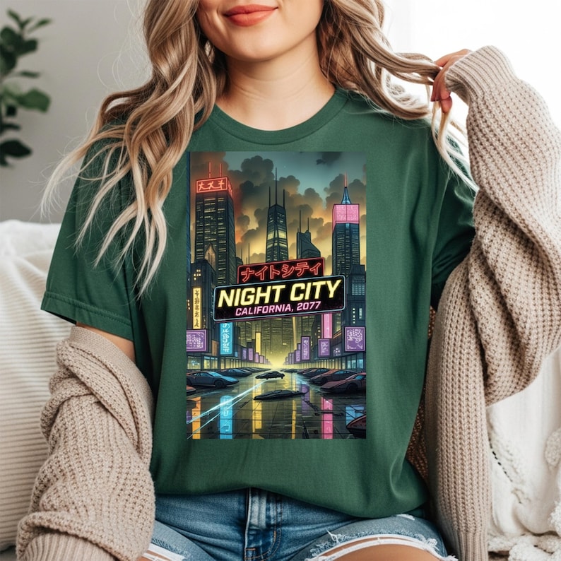 Op de afbeelding: Groen t-shirt met een afbeelding van een futuristisch stadslandschap met neonlichten en Japanse tekst. Het ontwerp bevat de woorden "NIGHT CITY CALIFORNIA, 2077". De illustratie toont hoge gebouwen en een reflecterend wateroppervlak.