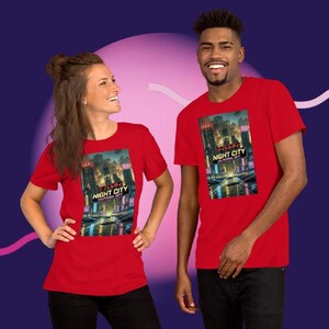 Op de afbeelding: Twee mensen dragen rode t-shirts met een afbeelding van een nachtelijk stadslandschap. De afbeelding bevat de tekst "NIGHT CITY CALIFORNIA CITY" in het wit en Japanse karakters. De shirts zijn korte mouwen.