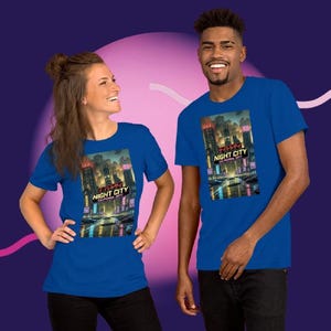 Op de afbeelding: Twee mensen dragen blauwe t-shirts met een afbeelding van een futuristisch stadslandschap 's nachts. De afbeelding bevat de tekst "NIGHT CITY CALIFORNIA 2077" in wit en rood. De shirts zijn korte mouwen.