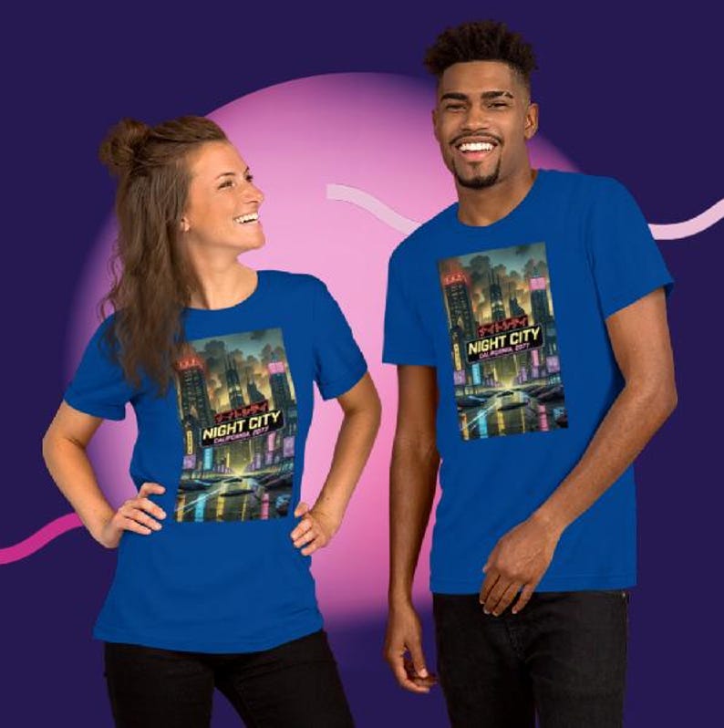 Op de afbeelding: Twee mensen dragen blauwe t-shirts met een afbeelding van een futuristisch stadslandschap 's nachts. De afbeelding bevat de tekst "NIGHT CITY CALIFORNIA 2077" in wit en rood. De shirts zijn korte mouwen.