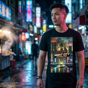 Op de afbeelding: Zwart t-shirt met een kleurrijke afbeelding van een futuristische stad 's nachts, met neonlichten. Het ontwerp bevat de tekst "NIGHT CITY CALIFORNIA, 2077" in het Engels en Japans. Een persoon draagt het t-shirt.