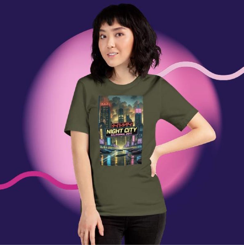 Op de afbeelding: Olijfgroen t-shirt met een levendige afbeelding van een futuristische stad, met de tekst "NIGHT CITY CALIFORNIA 2077". Het ontwerp toont neonlichten en hoge gebouwen, wat een cyberpunk esthetiek cre&euml;ert.