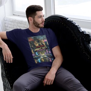 Op de afbeelding: Marineblauw T-shirt met een kleurrijke afbeelding van een nachtstad, met de tekst "Night City California 2077". De persoon draagt het T-shirt en zit op een zwarte chaise longue.