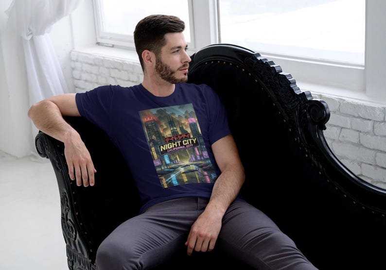Op de afbeelding: Marineblauw T-shirt met een kleurrijke afbeelding van een nachtstad, met de tekst "Night City California 2077". De persoon draagt het T-shirt en zit op een zwarte chaise longue.