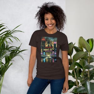 Op de afbeelding: Een bruin t-shirt met een levendige afbeelding van een futuristische stadsgezicht 's nachts. Het ontwerp bevat neonlichten, wolkenkrabbers en de tekst "NIGHT CITY CALIFORNIA 2077". Het shirt is gemaakt van een zacht materiaal.