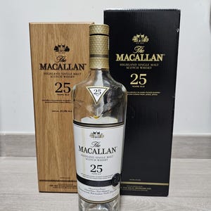 Puede incluir: Una botella de vidrio transparente de The Macallan Highland Single Malt Scotch Whisky, 25 años, con tapón dorado. La botella está flanqueada por una caja de madera y una caja negra, ambas con el logotipo y texto de la marca.