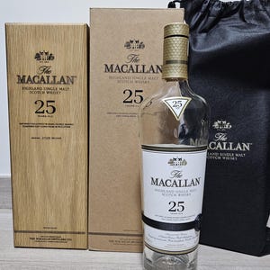 Puede incluir: Una botella de The Macallan 25 Years Old Highland Single Malt Scotch Whisky, con su caja de madera original y una caja de cartón. La botella tiene una tapa dorada y una etiqueta negra con texto blanco. También hay una bolsa negra.