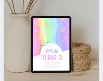 May include: A digital tablet shows a birthday invitation with a rainbow design and the text "AMELIA TURNS 5!". The tablet is on a wooden surface, next to a cream vase with dried stems and a small woven basket.