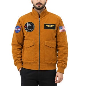 Könnte beinhalten: Eine senfgelbe Bomberjacke mit Stehkragen und durchgehendem Frontreißverschluss. Die Jacke ist mit einem NASA-Aufnäher, einem runden Missionsabzeichen, einem rechteckigen geflügelten Emblem und einer US-Flagge auf den Ärmeln und der Brust versehen.