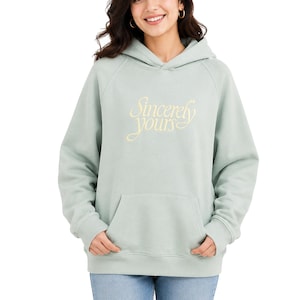 Könnte beinhalten: Ein langärmeliger Kapuzenpullover in Salbeigrün mit einer Kängurutasche auf der Vorderseite und dem Schriftzug "Sincerely yours" in hellgelber Schreibschrift auf der Brust. Das Kleidungsstück hat gerippte Bündchen und einen gerippten Saum.