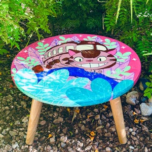 May include: A round, pink-topped wooden side table featuring a cartoon cat and bus design. The table has three wooden legs and is decorated with blue and green leaf-like shapes. The cat has a large grin.