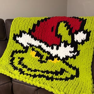 May include: A lime green, hand-knitted blanket featuring a cartoon face design with a red hat, black outline, white teeth, and yellow eyes. The blanket is made of a thick, soft yarn and is draped over a brown leather couch.