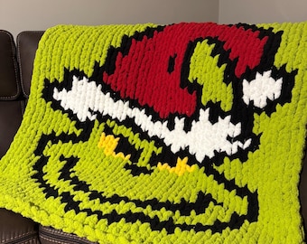 Puede incluir: Una manta de punto a mano de color verde lima con un diseño de cara de dibujos animados con un gorro rojo, contorno negro, dientes blancos y ojos amarillos. La manta está hecha de un hilo grueso y suave y está colocada sobre un sofá de cuero marrón.