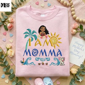 A pink t-shirt with a design featuring a cartoon character and the text "I am Momma" is displayed on a wooden surface, surrounded by flowers, beads, and a note.