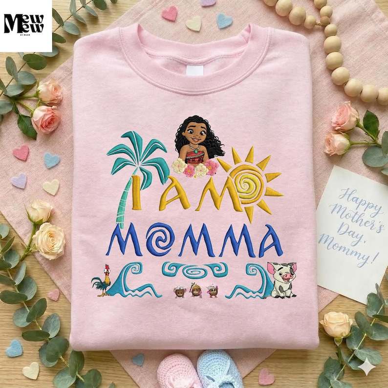 A pink t-shirt with a design featuring a cartoon character and the text "I am Momma" is displayed on a wooden surface, surrounded by flowers, beads, and a note.