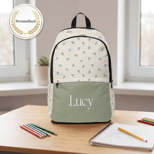 May include: A backpack featuring a cream-colored upper section with a light blue strawberry pattern and a solid sage green lower pocket. The name Lucy is printed in white text on the front pocket. A circular badge reads Personalized.