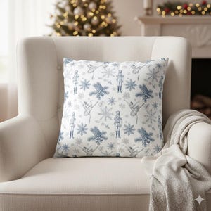 May include: A square decorative pillow with a white background and a blue pattern featuring snowflakes, ballerinas, and nutcrackers. The pillow is placed on a cream-colored armchair, with a gray blanket draped over the arm. A Christmas tree is visible in the background.