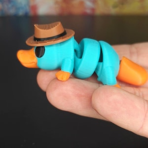 以下が含まれることがあります： ターコイズ、オレンジ、茶色の3Dプリントされた関節式カモノハシのおもちゃ。おもちゃは茶色のフェドラハット、オレンジ色のくちばしと足、セグメント化された体を特徴としています。おもちゃは手で持たれています。