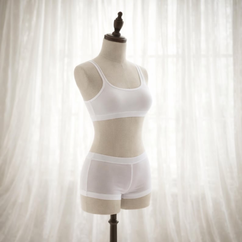 May include: A white lingerie set displayed on a mannequin. The set includes a simple, scoop-neck bralette and matching boy shorts. The fabric appears to be a soft, lightweight material. The mannequin is set against a backdrop of sheer white curtains.
