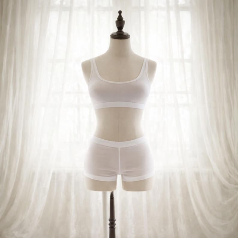 May include: A white lingerie set displayed on a mannequin. The set includes a scoop-neck bralette and matching shorts. The fabric appears lightweight and semi-sheer. The mannequin is set against a backdrop of a window with white curtains.
