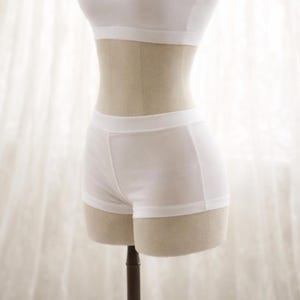 May include: A white two-piece lingerie set displayed on a mannequin. The set includes a bandeau-style top and a pair of boy-short style bottoms. The fabric appears to be a soft, lightweight material. The background is a sheer white curtain.