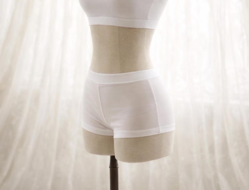 May include: A white two-piece lingerie set displayed on a mannequin. The set includes a bandeau-style top and a pair of boy-short style bottoms. The fabric appears to be a soft, lightweight material. The background is a sheer white curtain.