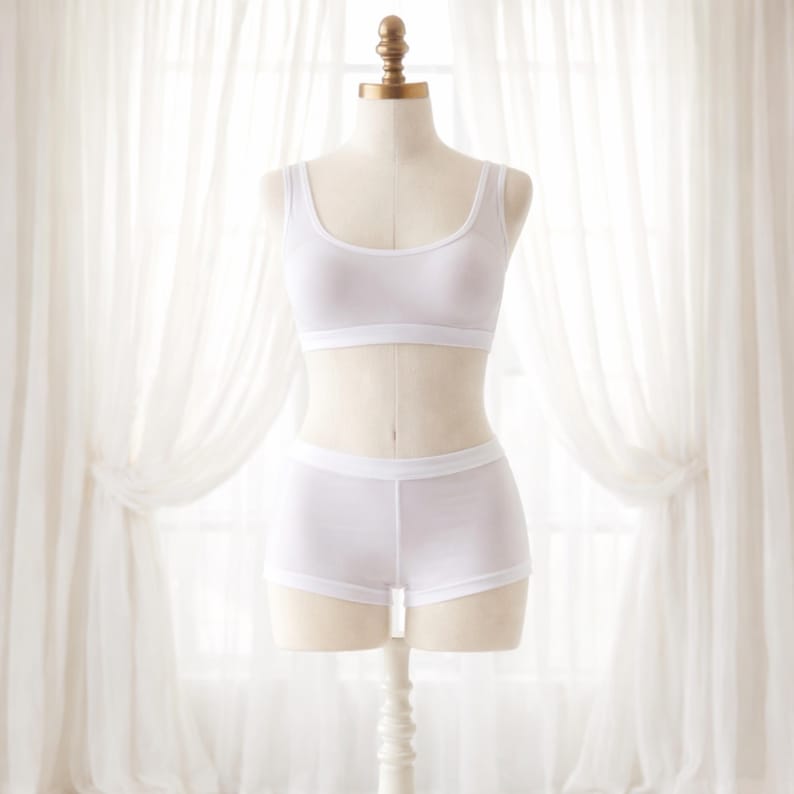 May include: A white lingerie set displayed on a mannequin. The set includes a scoop-neck sports bra and matching boy shorts. The fabric appears to be a soft, lightweight material. The mannequin is positioned in front of a window with sheer white curtains.