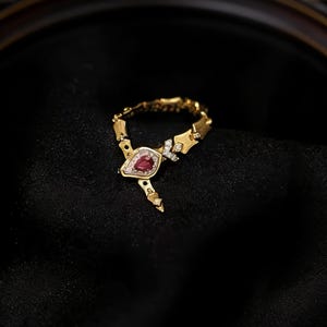 May include: A gold-toned ring with a teardrop-shaped, red gemstone center. The ring features a delicate design with small diamonds and is set against a black background. The jewelry piece is an elegant accessory.