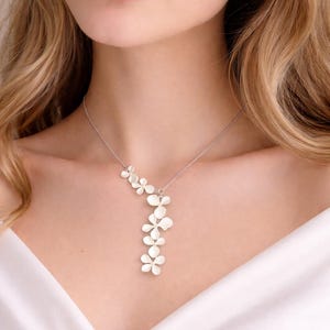 Puede incluir: Un collar plateado con una cascada de colgantes de flores blancas. El delicado diseño floral cuelga de una fina cadena, creando un accesorio elegante y femenino. El collar se muestra sobre un fondo blanco.
