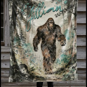 May include: A soft blanket featuring a watercolour-style illustration of a brown Bigfoot walking through a forest. The name Jillian is printed in large, teal cursive script at the top. The scene includes muted green and brown tones with trees.