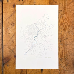 May include: A detailed map print of Bend, Oregon, featuring contour lines and a blue river. The print is on white paper with the city name and "566" printed in the bottom right corner. The map is a minimalist design.